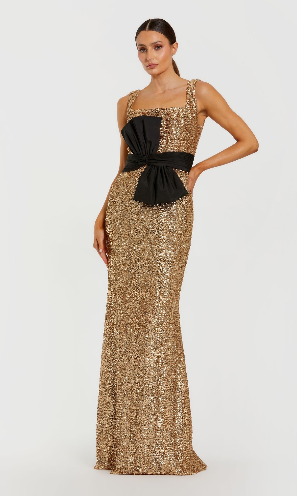 Woman wearing a gold sequin dress with a black bow detail at the waistline standing in front of a white background